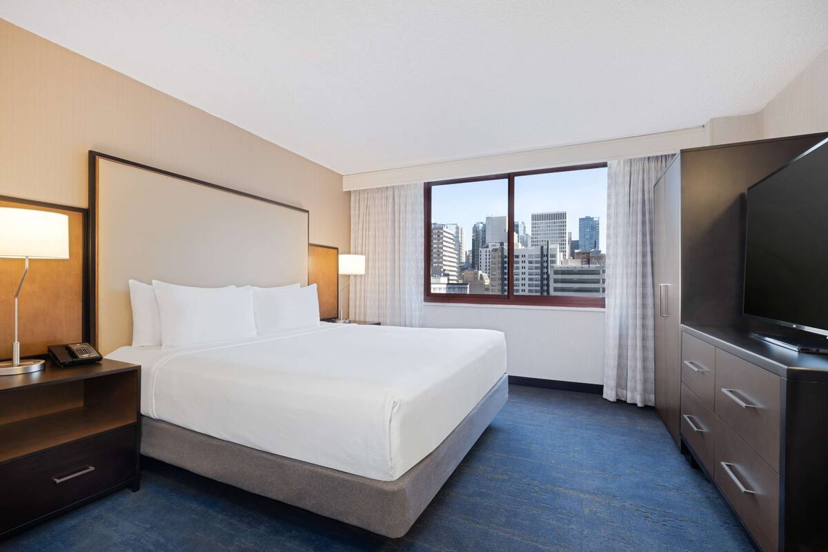 A hotel room with a large bed, a desk with a lamp, a TV, and a window with a city view.