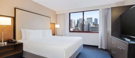 A hotel room with a large bed, a desk with a lamp, a TV, and a window with a city view.