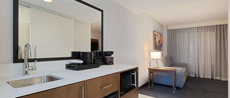 A modern hotel room with a kitchenette, a large mirror, a coffee machine, a small table with a lamp, and a view of the city through the window.