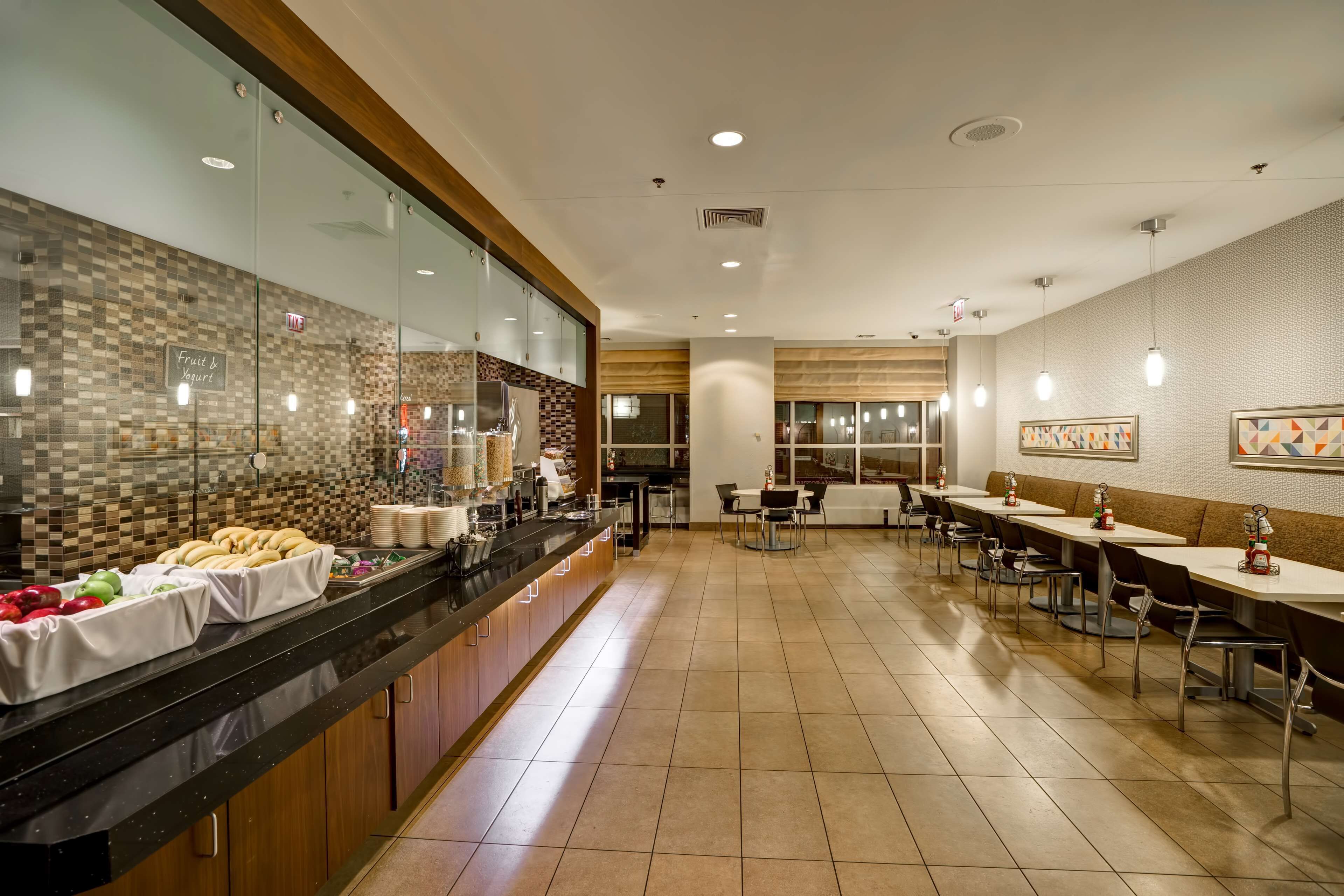 A modern restaurant with a long counter, glass partitions, and a variety of food items displayed. There are multiple tables and chairs arranged for dining.
