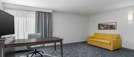 A hotel room with a desk, chair, sofa, and a painting on the wall.