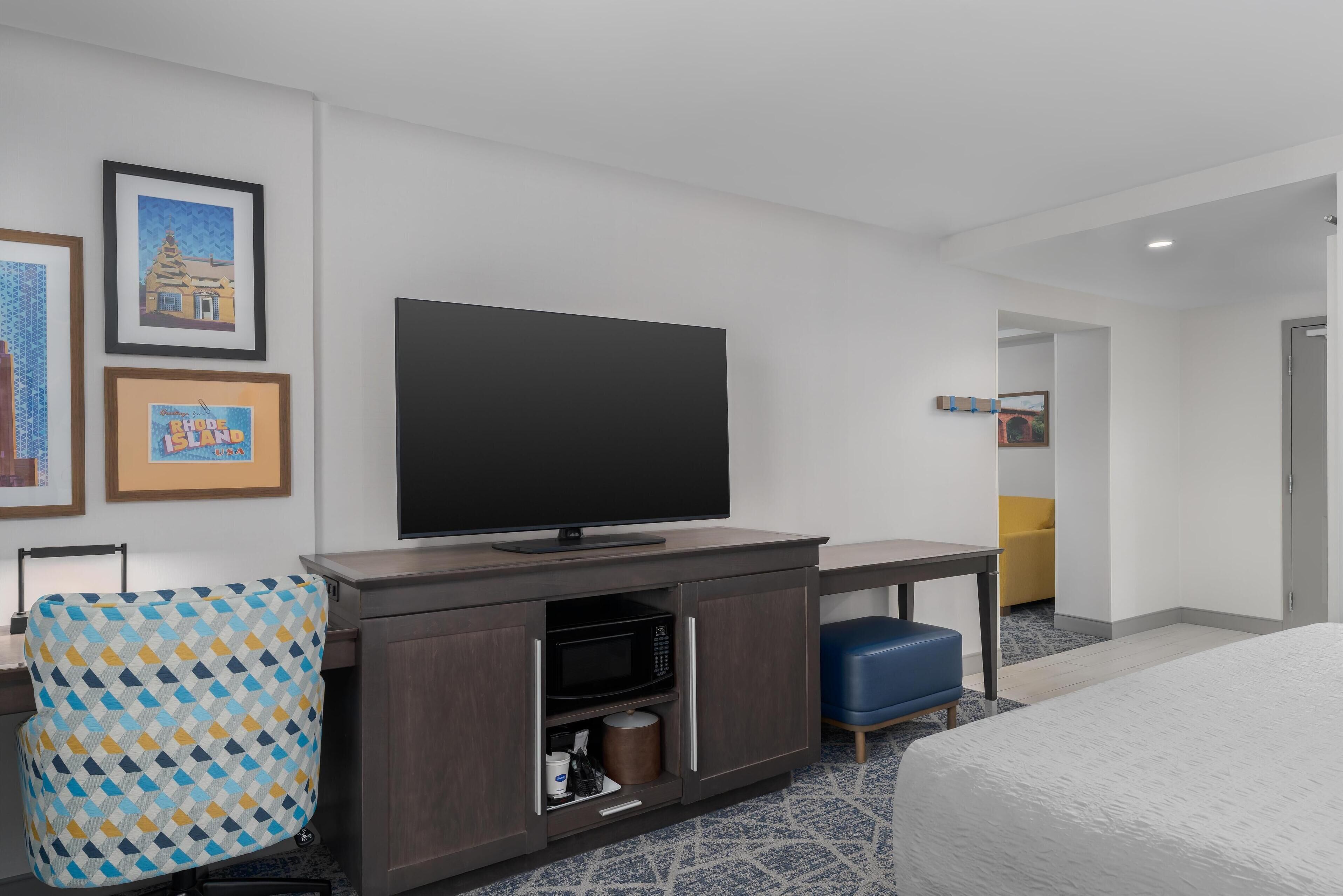 A hotel room with a flat-screen TV, a desk, a microwave, a blue chair, and a bed.