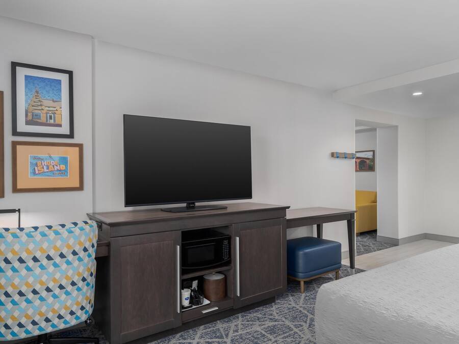 A hotel room with a flat-screen TV, a desk, a microwave, a blue chair, and a bed.