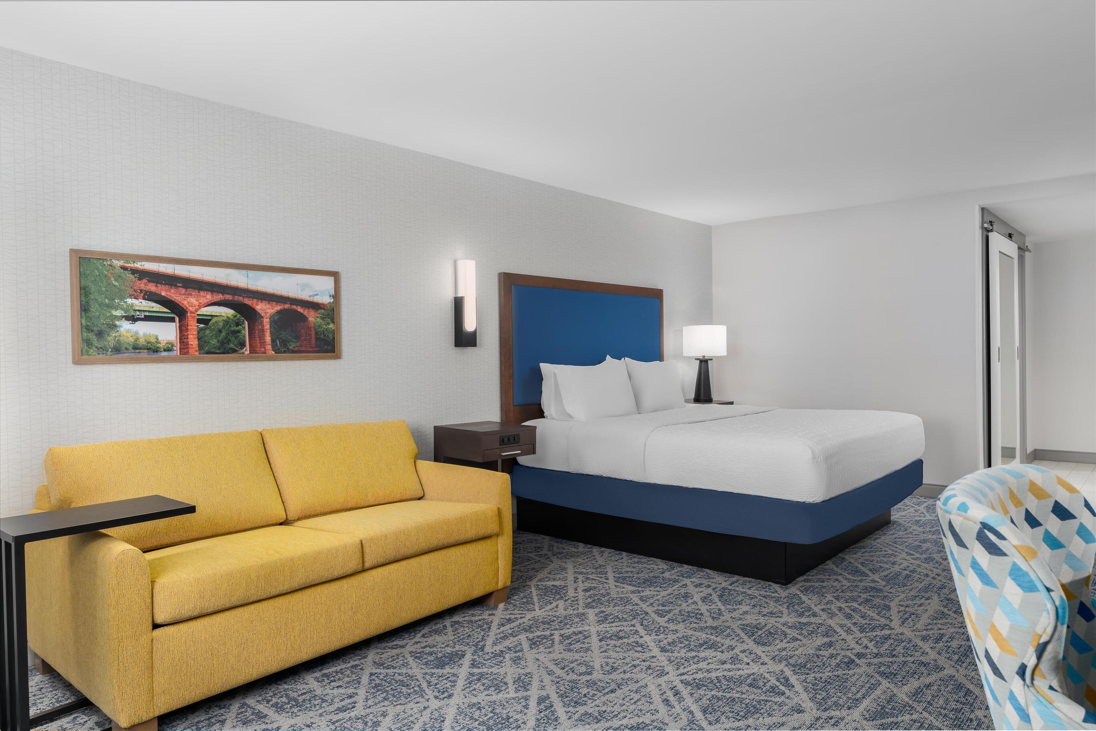 A hotel room with a bed, a sofa, a nightstand, a lamp, and a painting on the wall.
