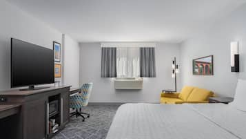 A hotel room with a bed, a desk with a flat-screen TV, a chair, a sofa, an air conditioning unit, and a window with curtains.