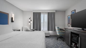 Ein Hotelzimmer mit einem Bett, einem Schreibtisch, einem Fernseher, einem Sessel und einem großen Fenster mit Vorhängen.