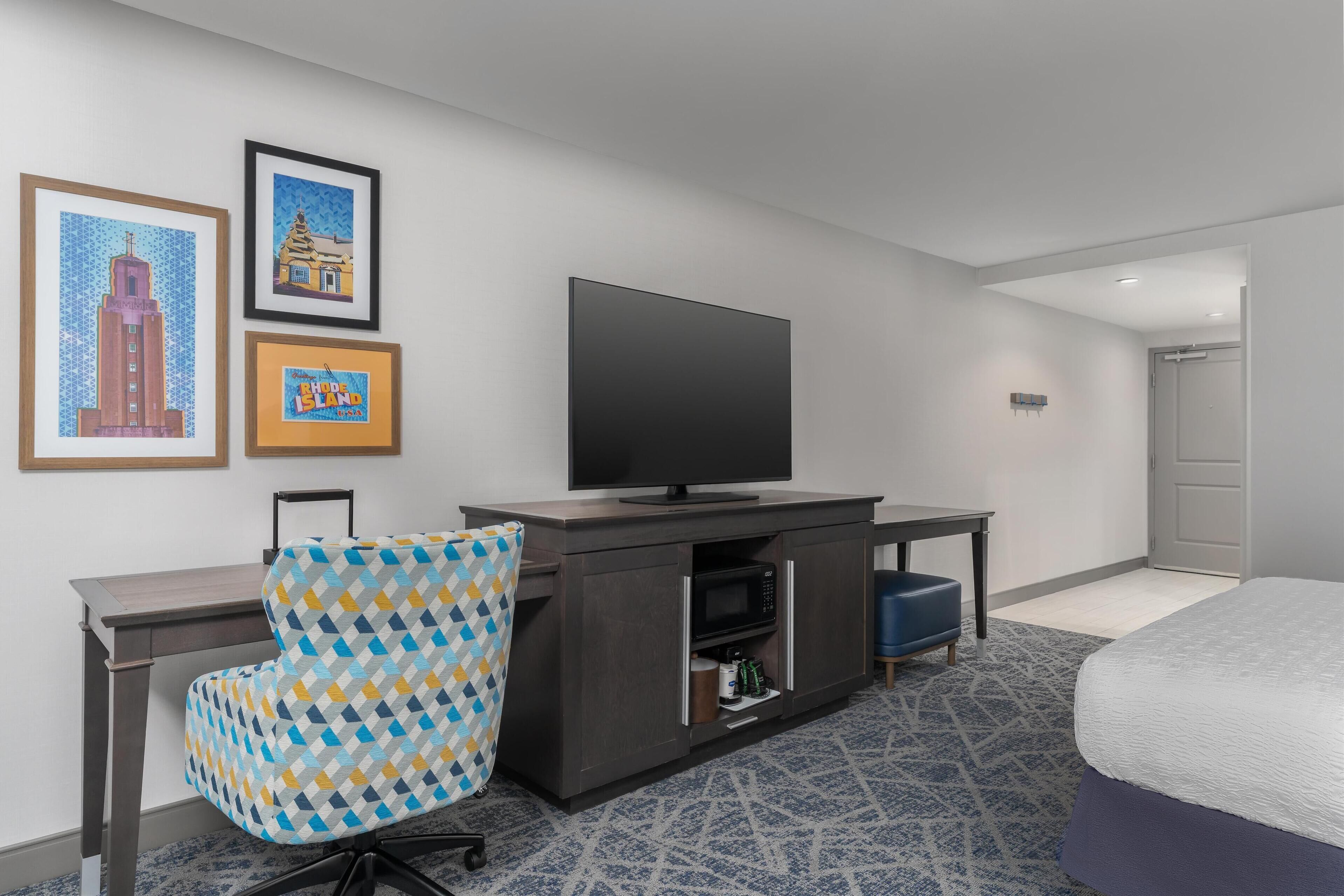 Habitación de hotel con cama, escritorio con televisor, sillón estampado y obra de arte en la pared.