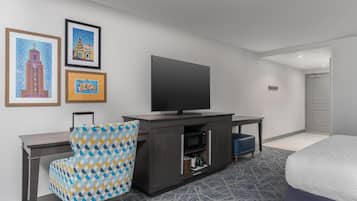 Habitación de hotel con cama, escritorio con televisor, sillón estampado y obra de arte en la pared.