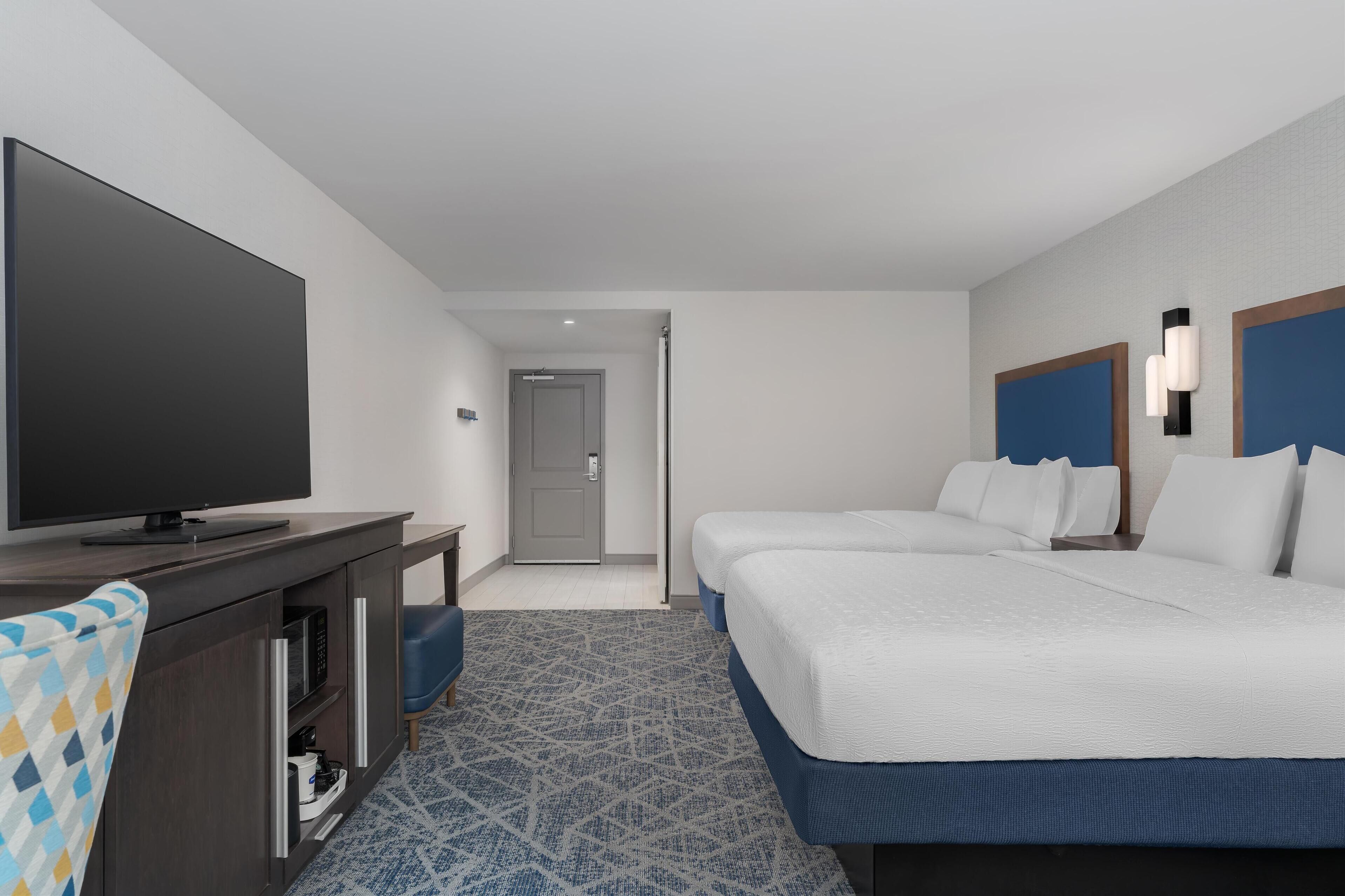 A hotel room with a flat-screen TV, a desk with a microwave, two beds with blue headboards, and a patterned carpet.