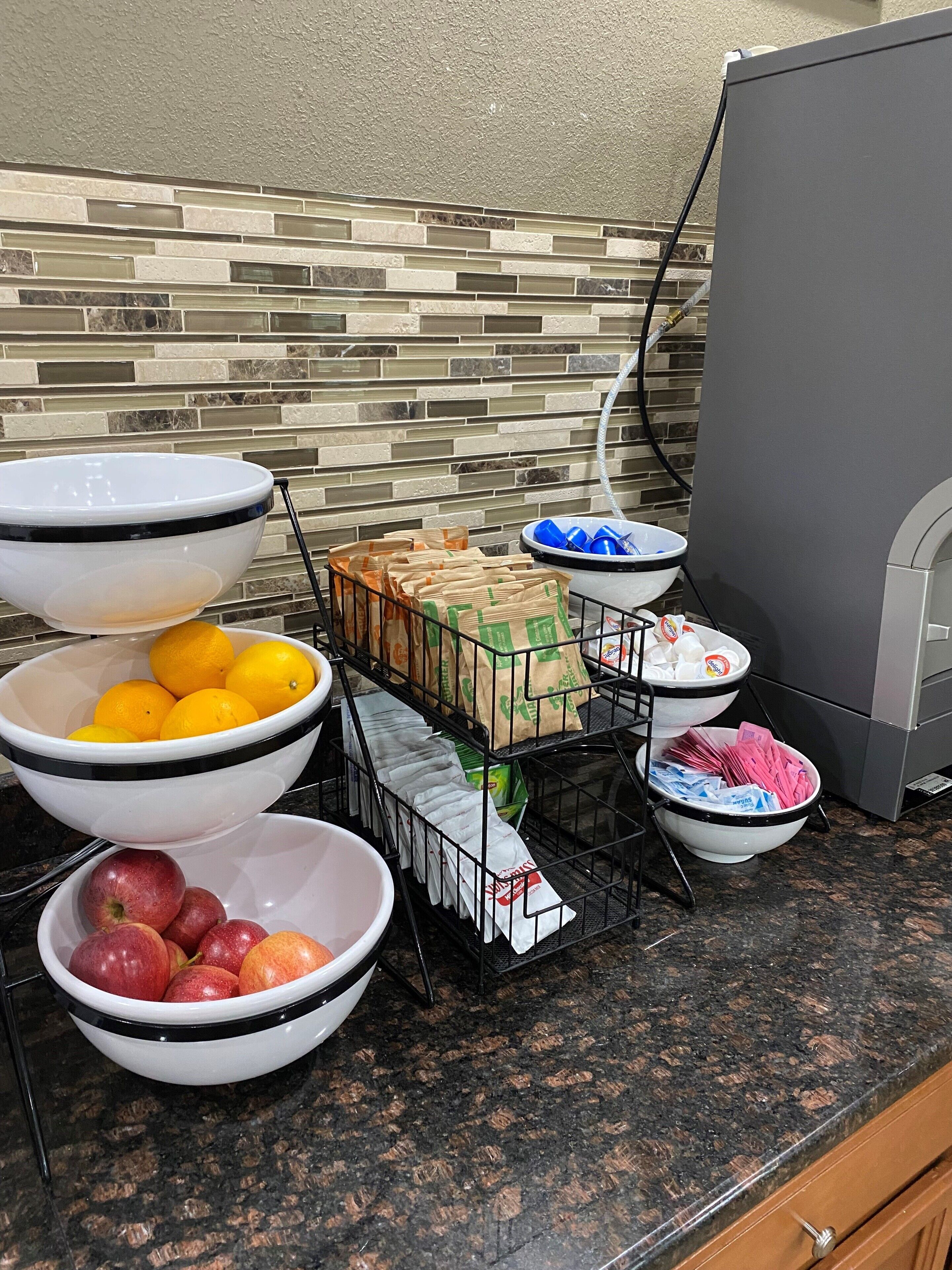 A kitchen counter with various bowls containing fruits and packaged goods, a coffee maker, and a tiled backsplash.
