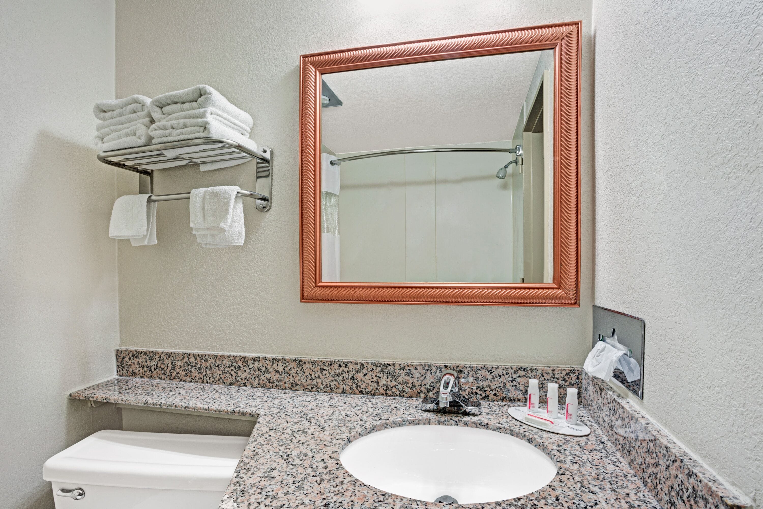 A bathroom with a granite countertop sink, a white toilet, a towel rack with folded towels, and a large mirror with a copper frame.