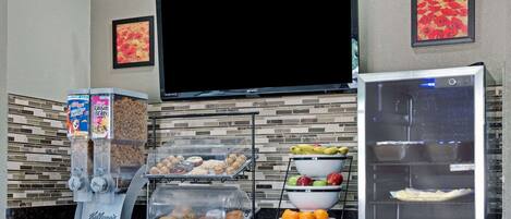 A breakfast bar with cereal dispensers, a microwave, and a refrigerator stocked with beverages and fruits.