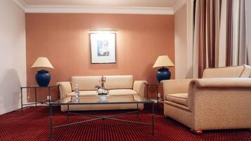 Habitación de hotel con sofá, sillón, mesa de centro de vidrio, dos lámparas, un cuadro en la pared y piso cubierto de alfombra roja.