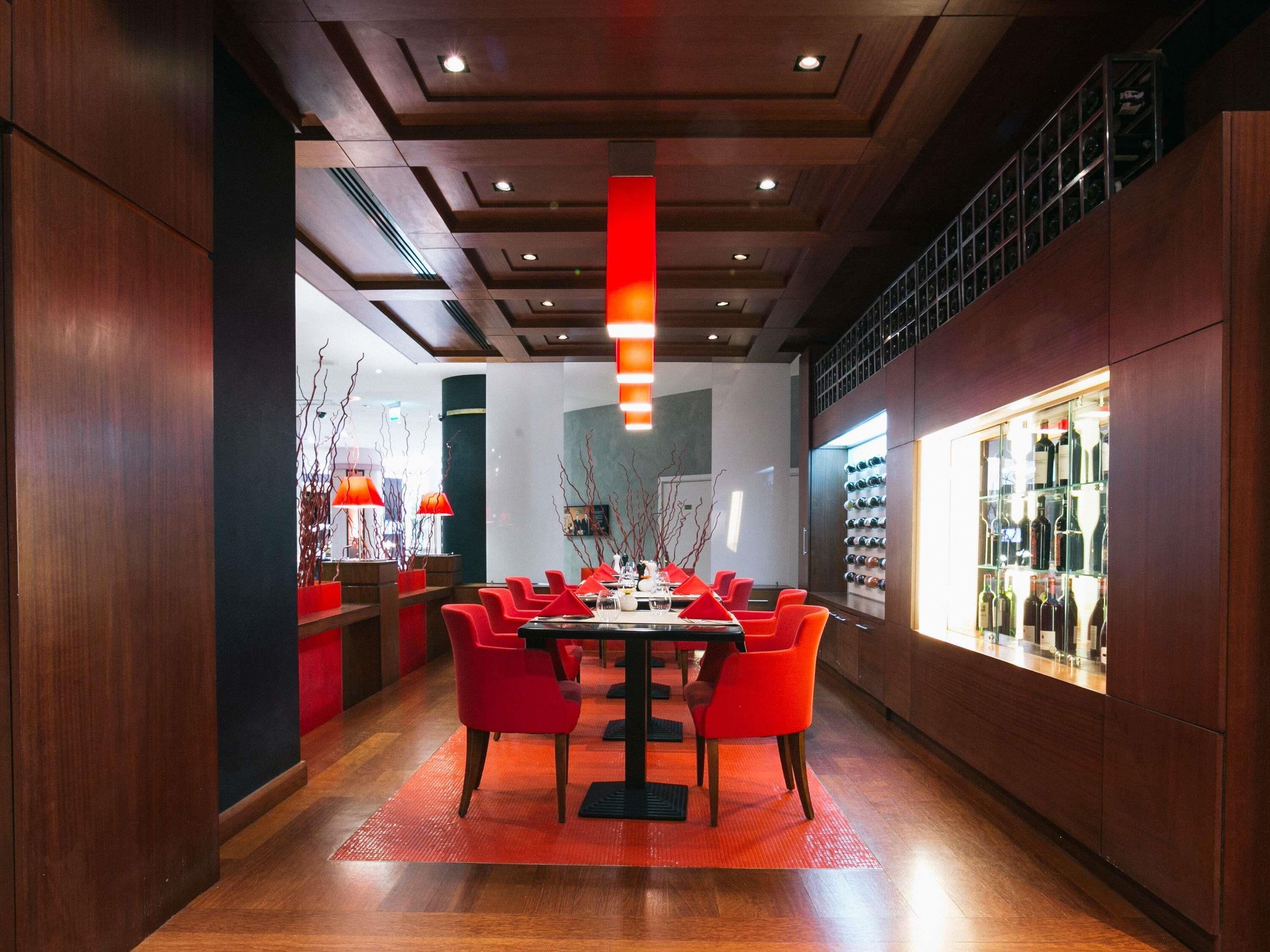 A modern dining area with a long table, red chairs, and a wine display on the right.