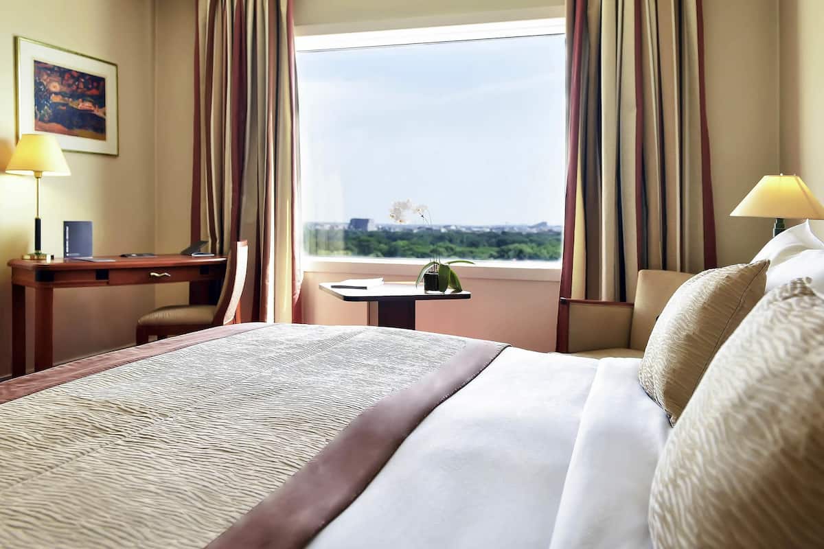 A hotel room with a bed, desk, chair, and a view of the cityscape through large windows.