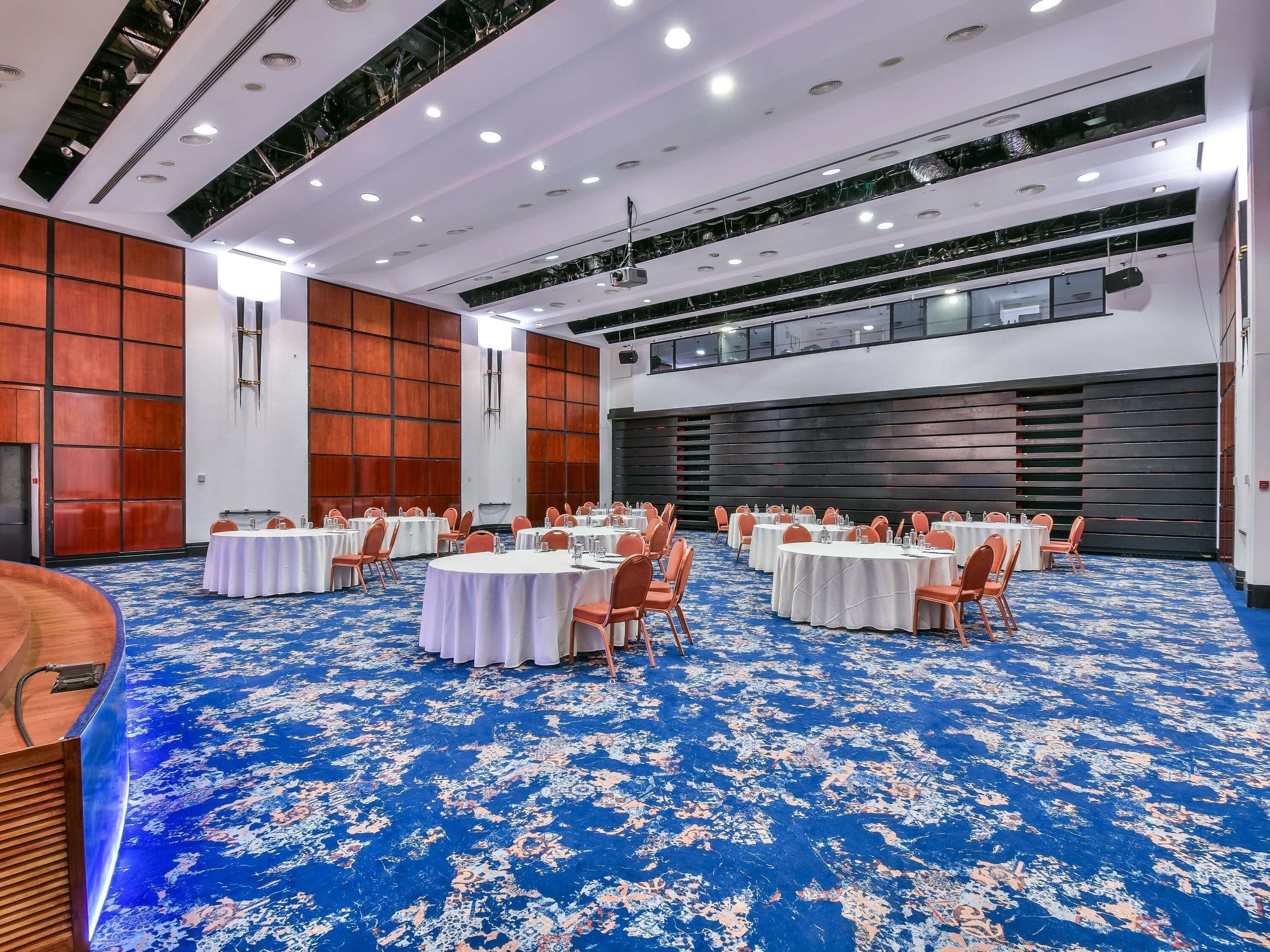 A large conference room with round tables, orange chairs, and a blue patterned carpet.