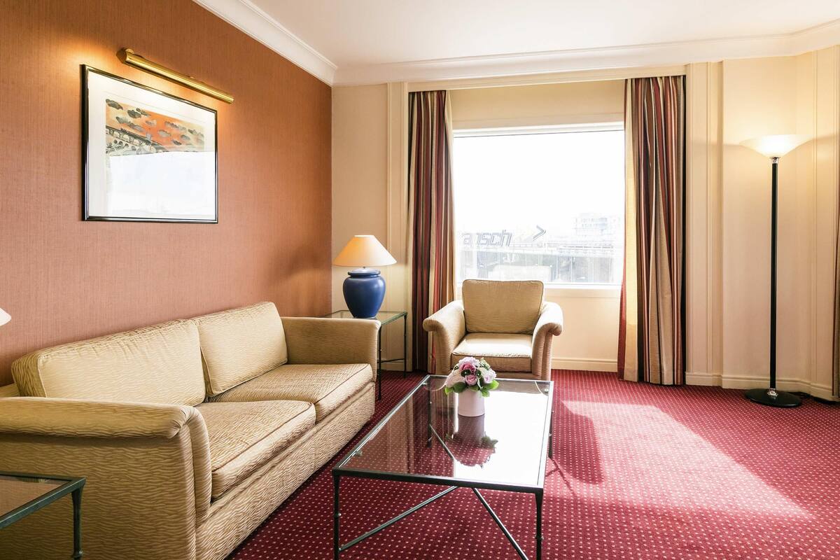 A hotel room with a beige sofa, a small table with a vase of flowers, a lamp, and a view of the city through large windows.