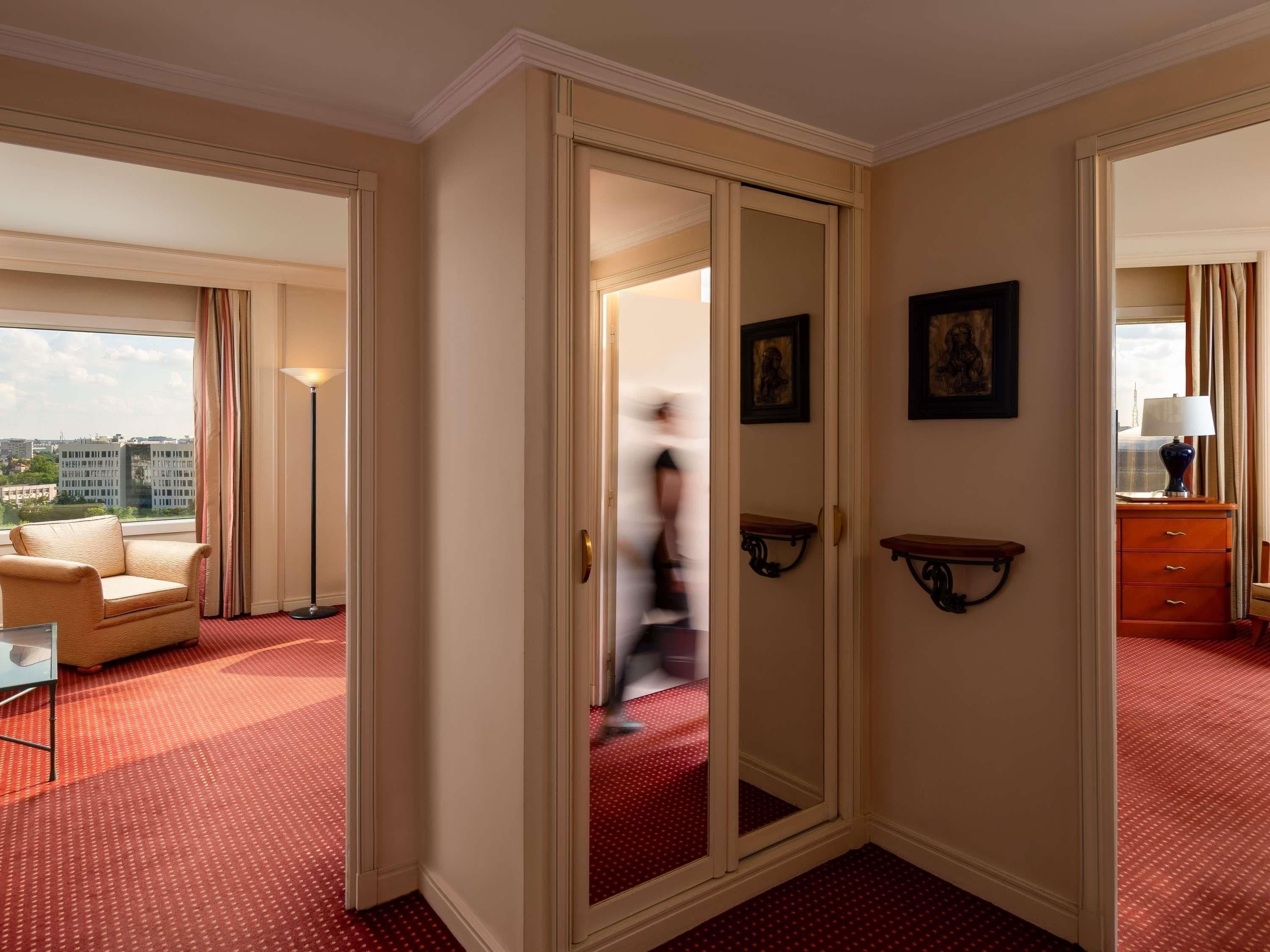 A hotel room with a large window, a red carpet, a sofa, a mirror, and a dresser.