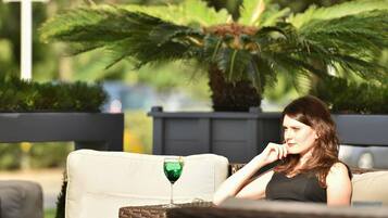 Una mujer sentada en un sofá de mimbre con una bebida verde, rodeada de follaje y un entorno moderno al aire libre.