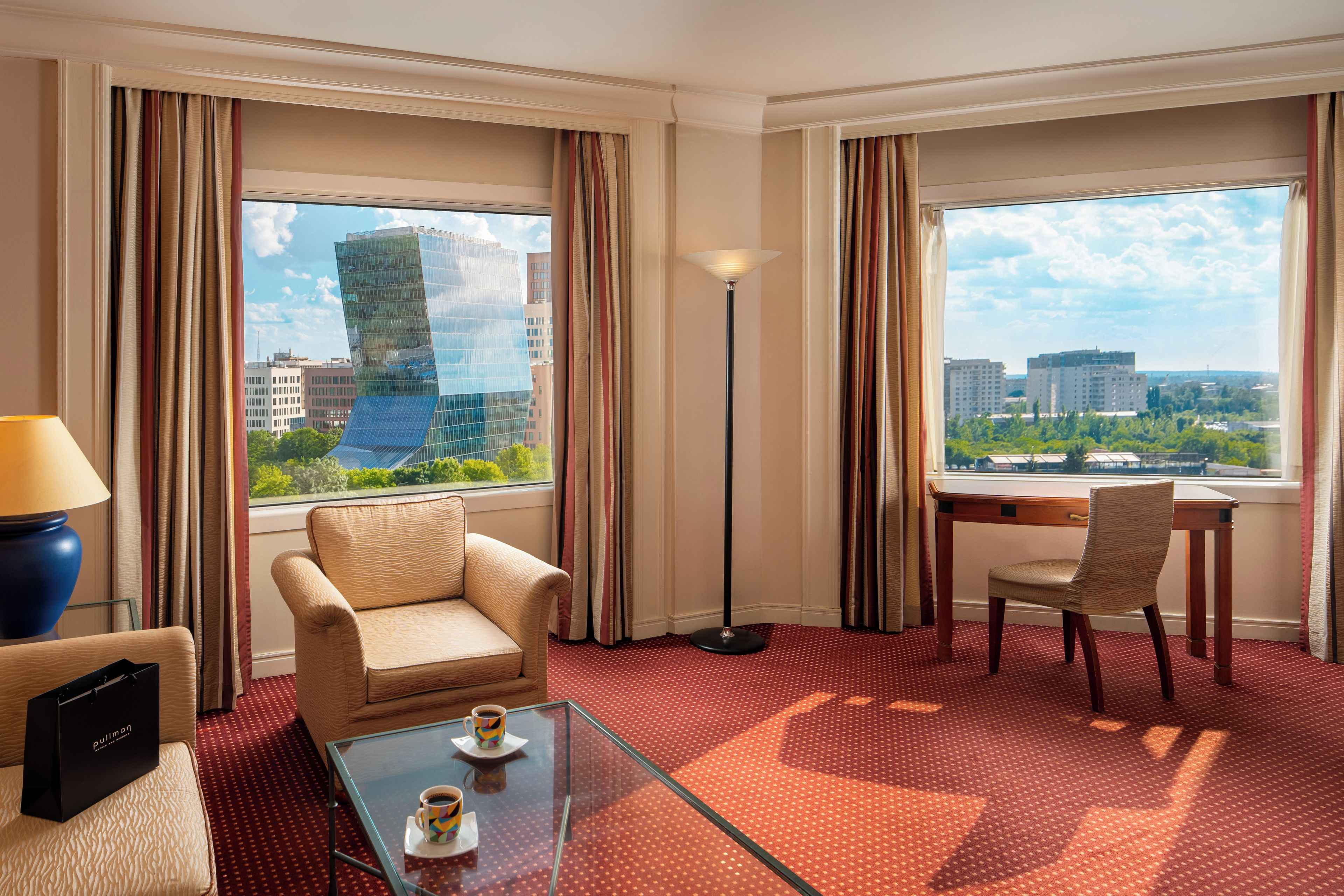 A hotel room with a sofa, a chair, a small table with a cup, a lamp, and a view of a cityscape through large windows.