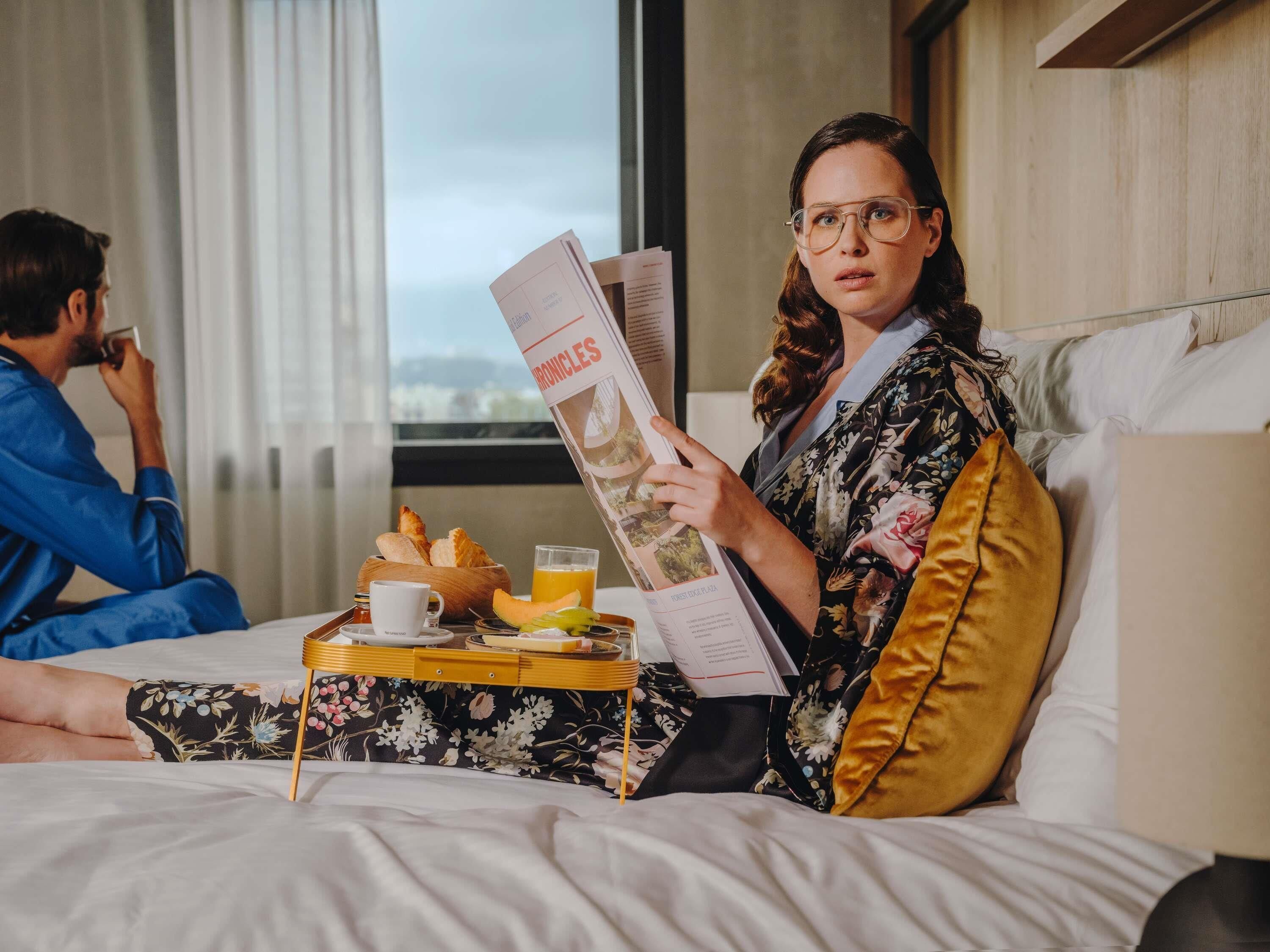 A person reading a newspaper on a bed with a tray of breakfast items.