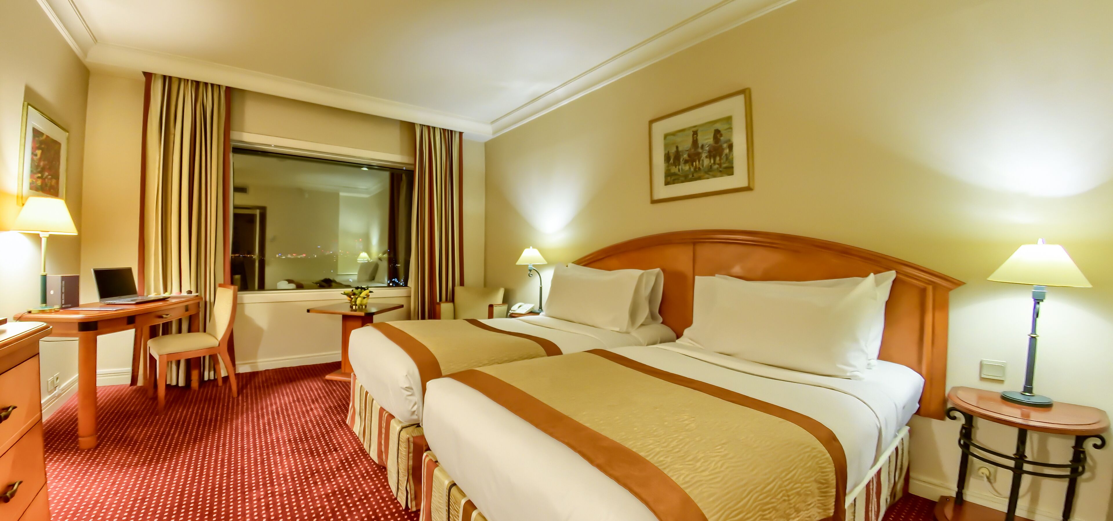 A hotel room with two beds, a desk, a chair, a lamp, and a painting on the wall.