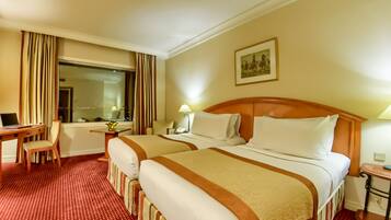 A hotel room with two beds, a desk, a chair, a lamp, and a painting on the wall.