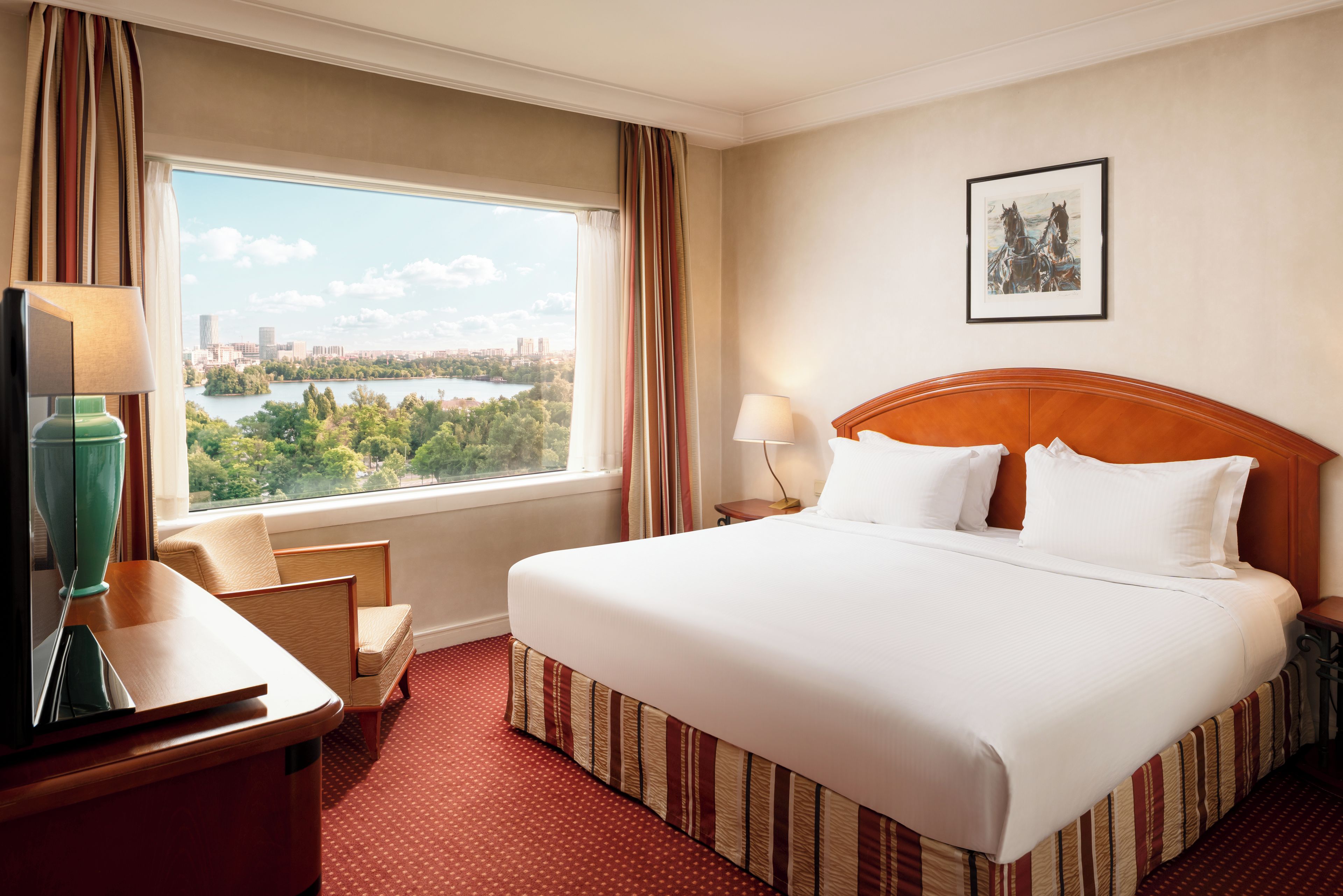 Una habitación de hotel con una cama grande, un escritorio con lámpara, una silla, un ventanal con vistas a la ciudad y un cuadro en la pared.