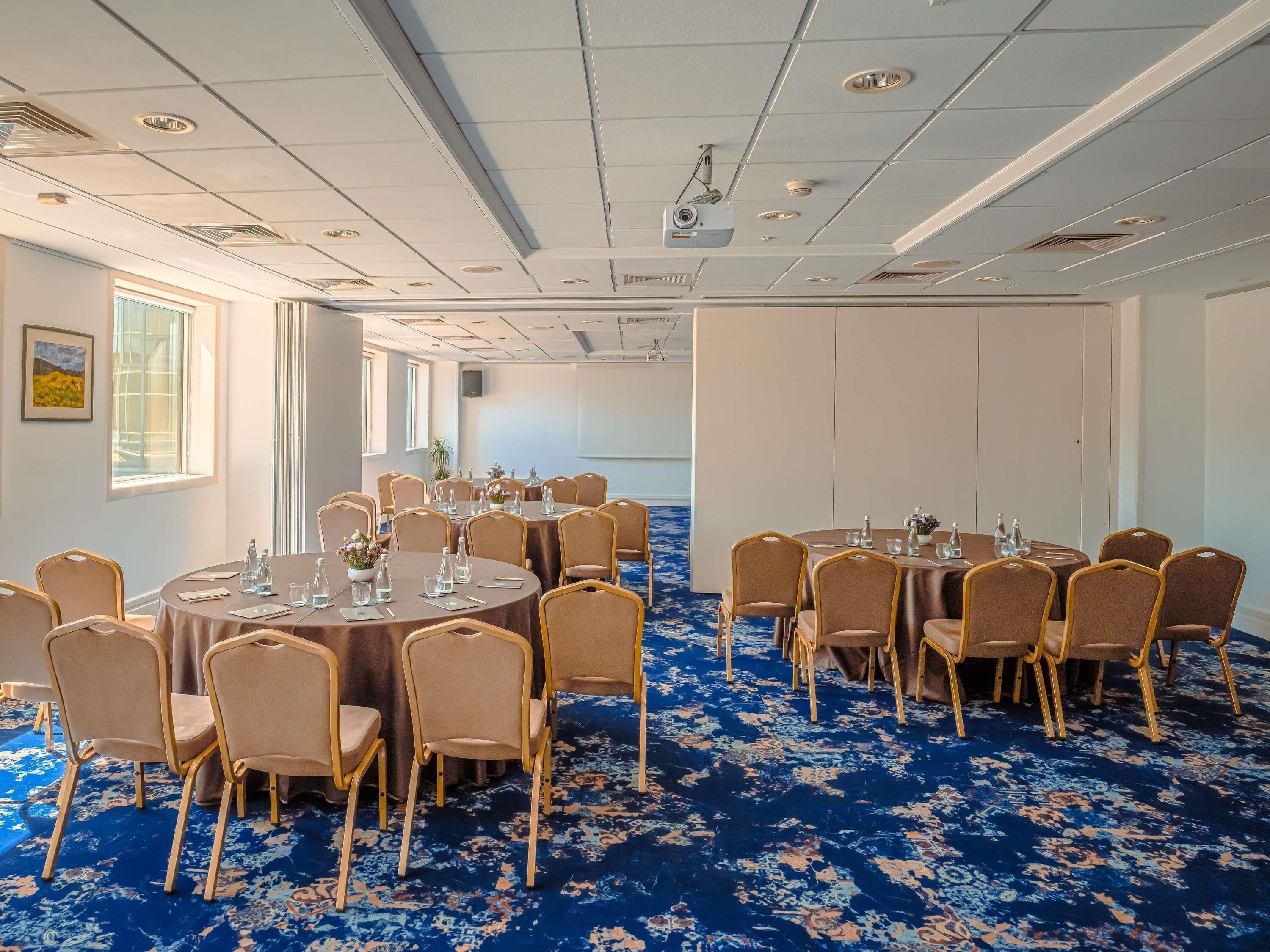 Un salón de conferencias con mesas redondas, sillas y un proyector en el techo.