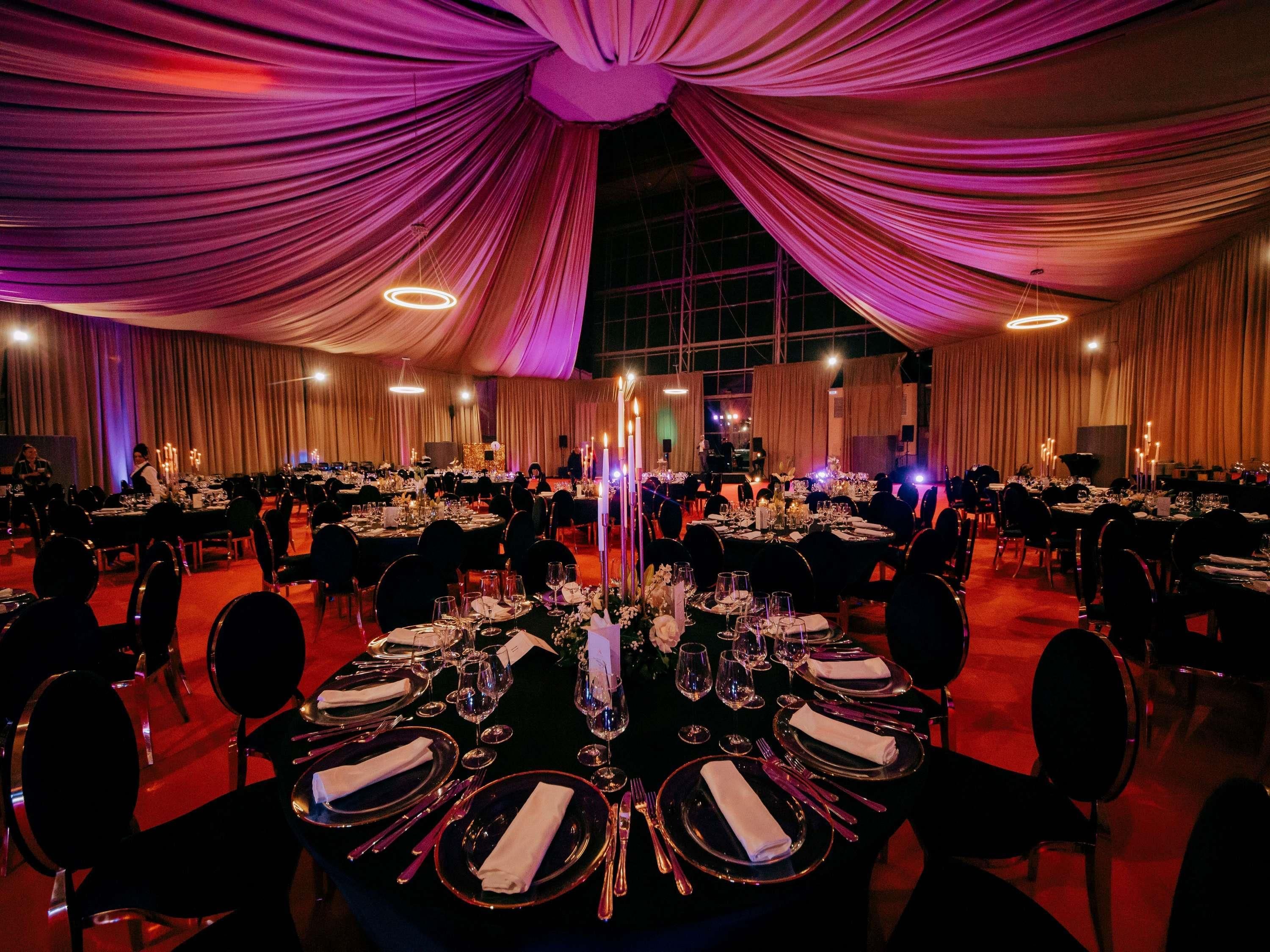 A banquet hall with round tables set for a formal event, draped with purple curtains, and equipped with candles and table settings.