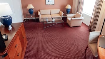 A hotel room with a red carpet, a sofa, a chair, a small table, a lamp, and a picture on the wall.