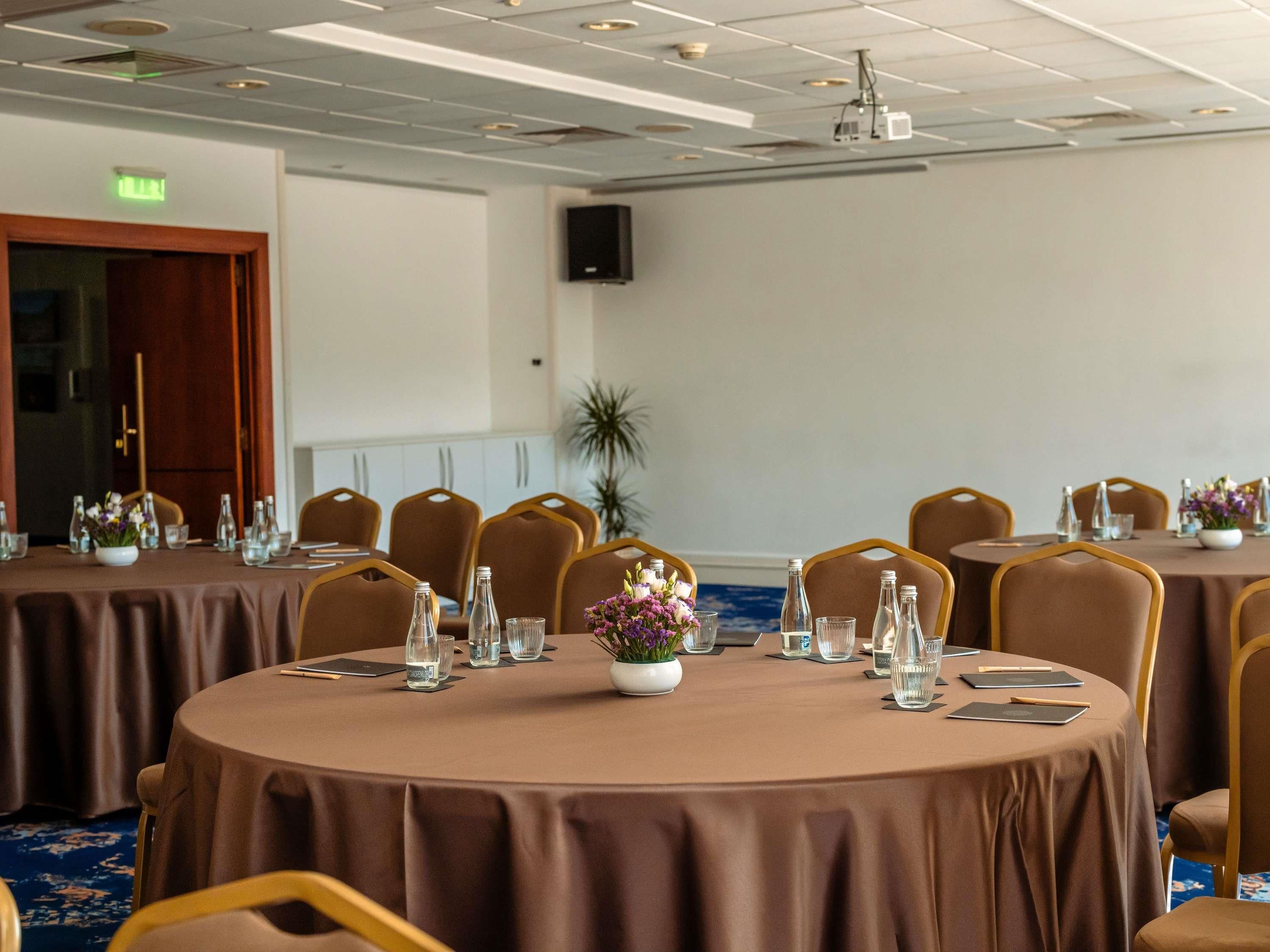 Un salón de conferencias con mesas redondas, sillas y un altavoz en la pared.