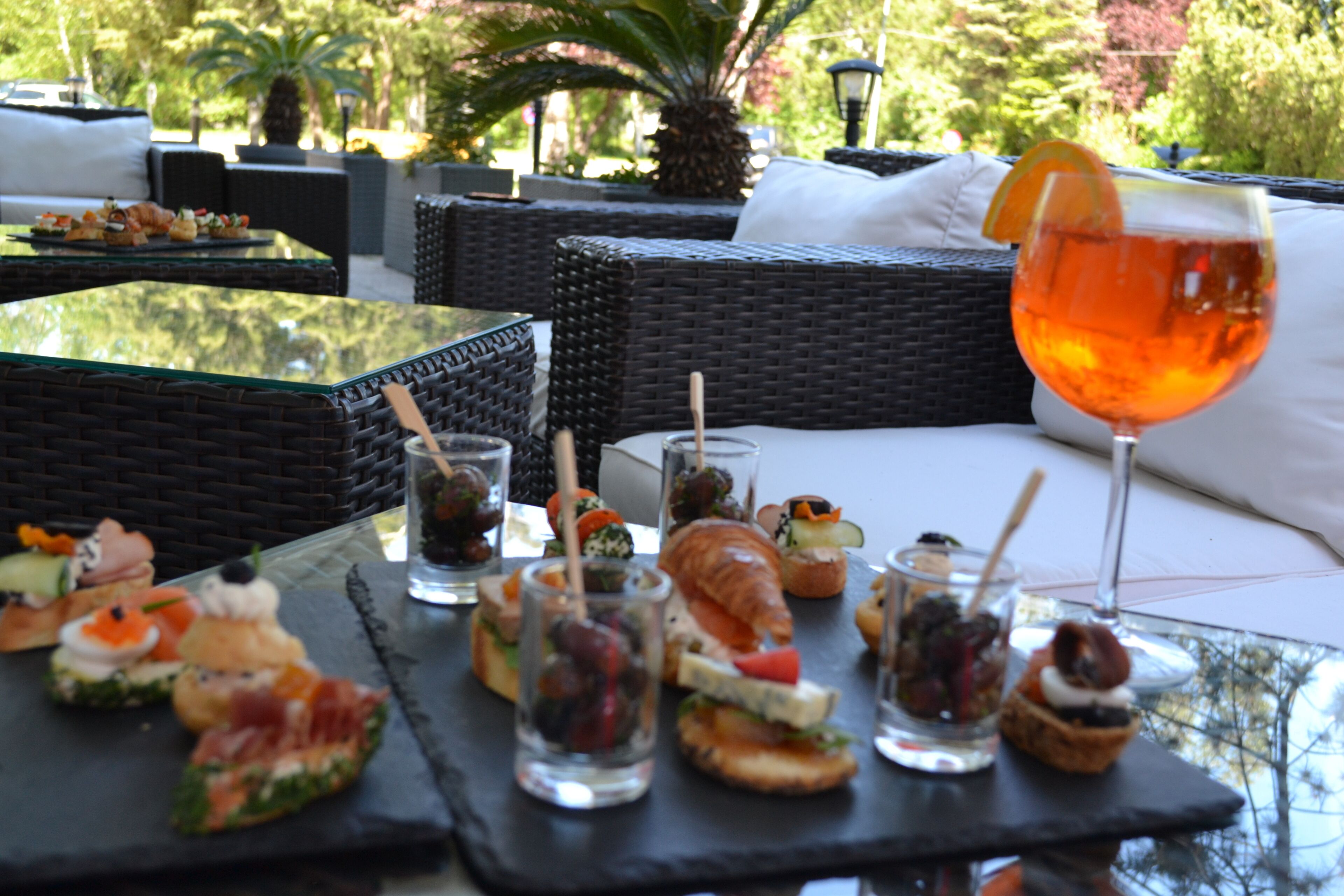 Una mesa con diferentes aperitivos y un vaso de jugo de naranja, ubicada en un patio exterior con mobiliario de mimbre y vistas a la vegetación.
