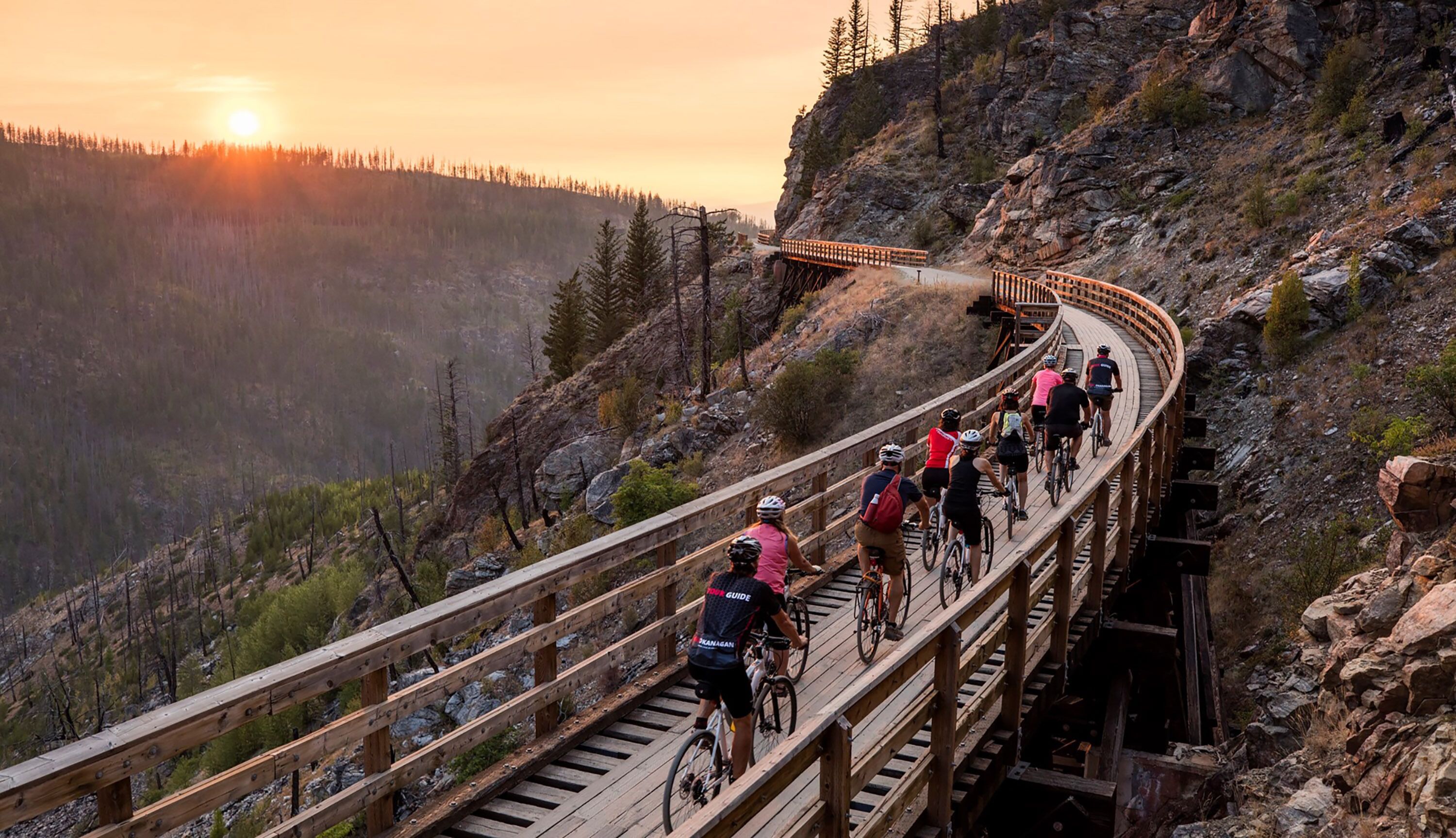 A wooden bridge with cyclists on it, surrounded by rocky terrain and trees.