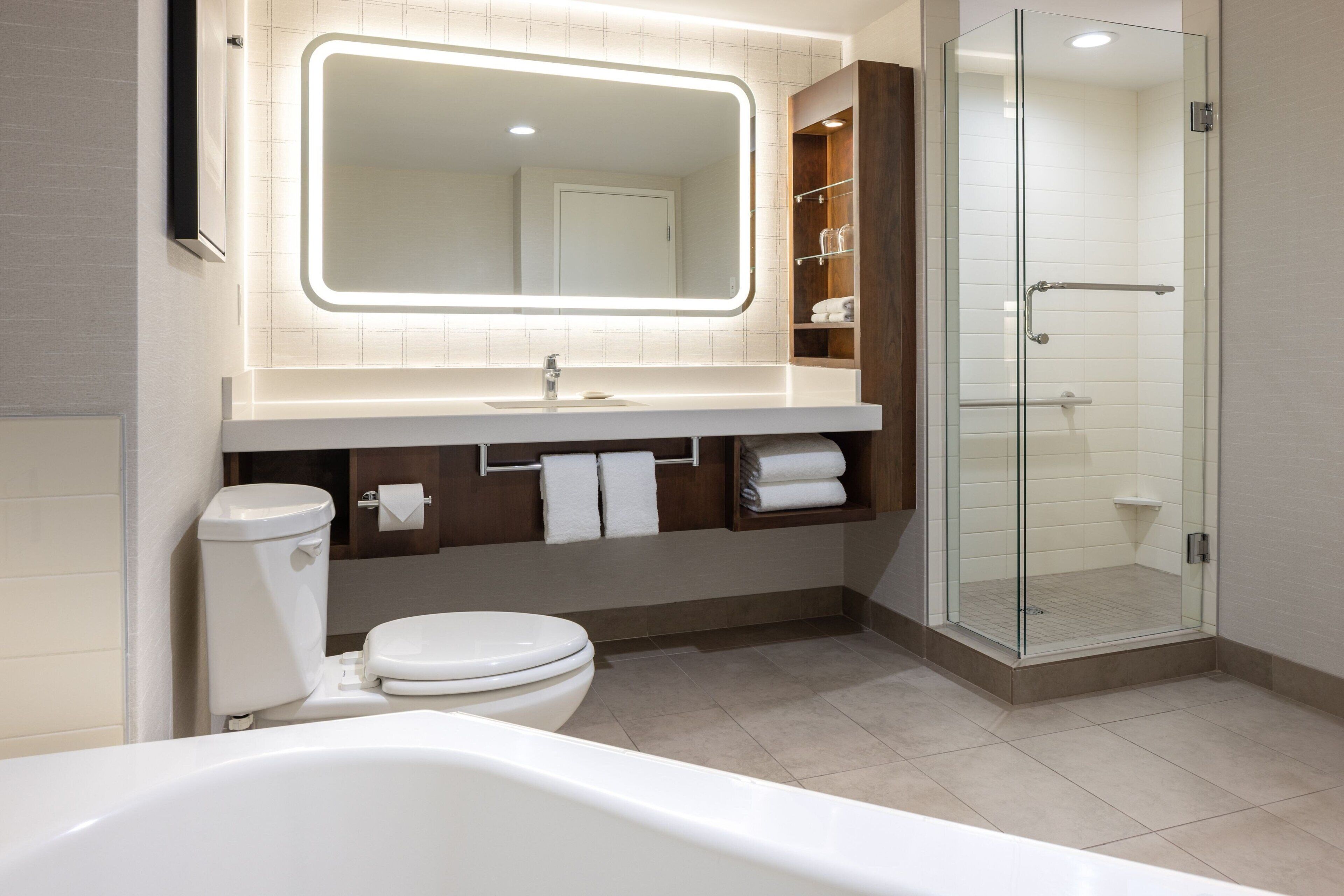 Une salle de bain moderne avec une douche à parois de verre, un grand miroir doté d’un éclairage indirect, des toilettes blanches et une baignoire.