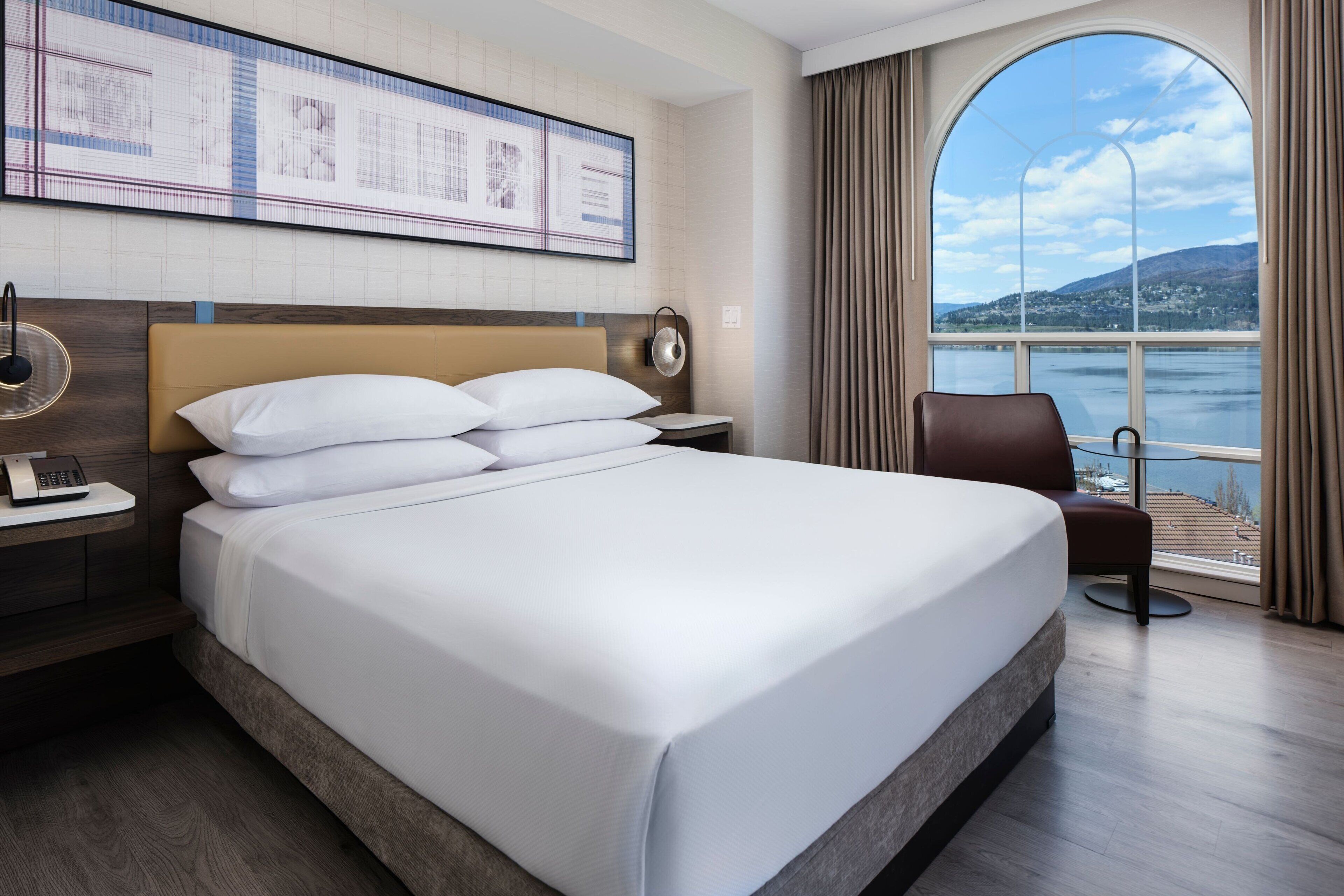 A hotel room with a large bed, a nightstand with a phone, a wall-mounted clock, and a large window with a view of a body of water and hills.