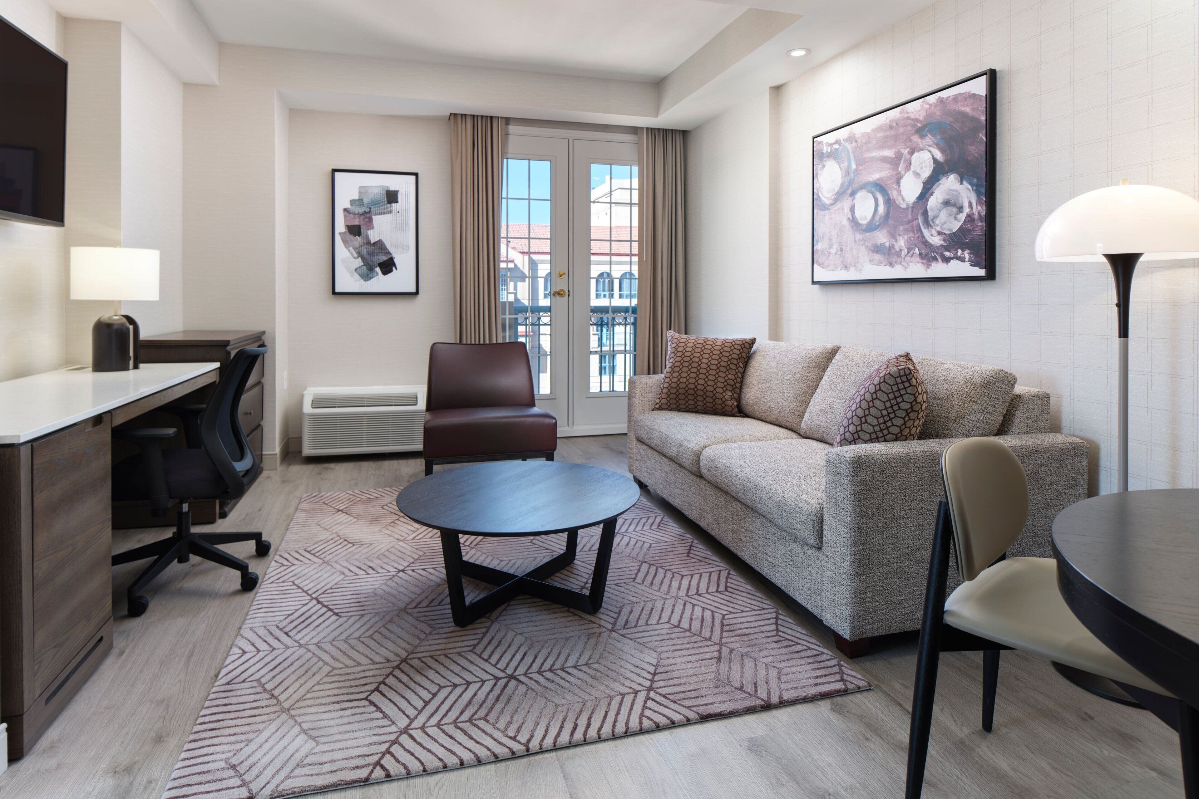 A modern hotel room with a sofa, a desk, a chair, a round coffee table, and a large window with a view of buildings.