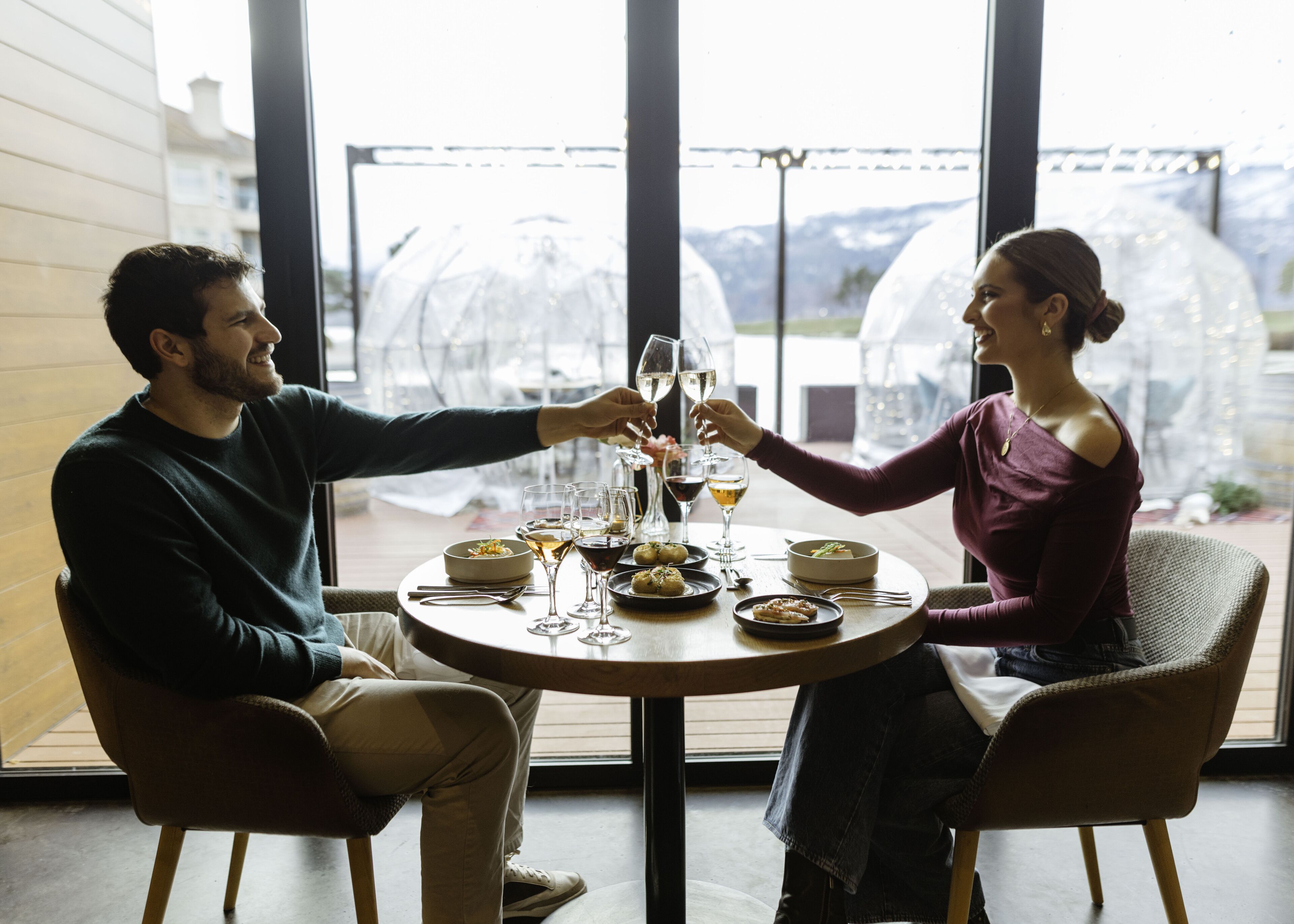A couple seated at a round table with a view of mountains through a glass enclosure.