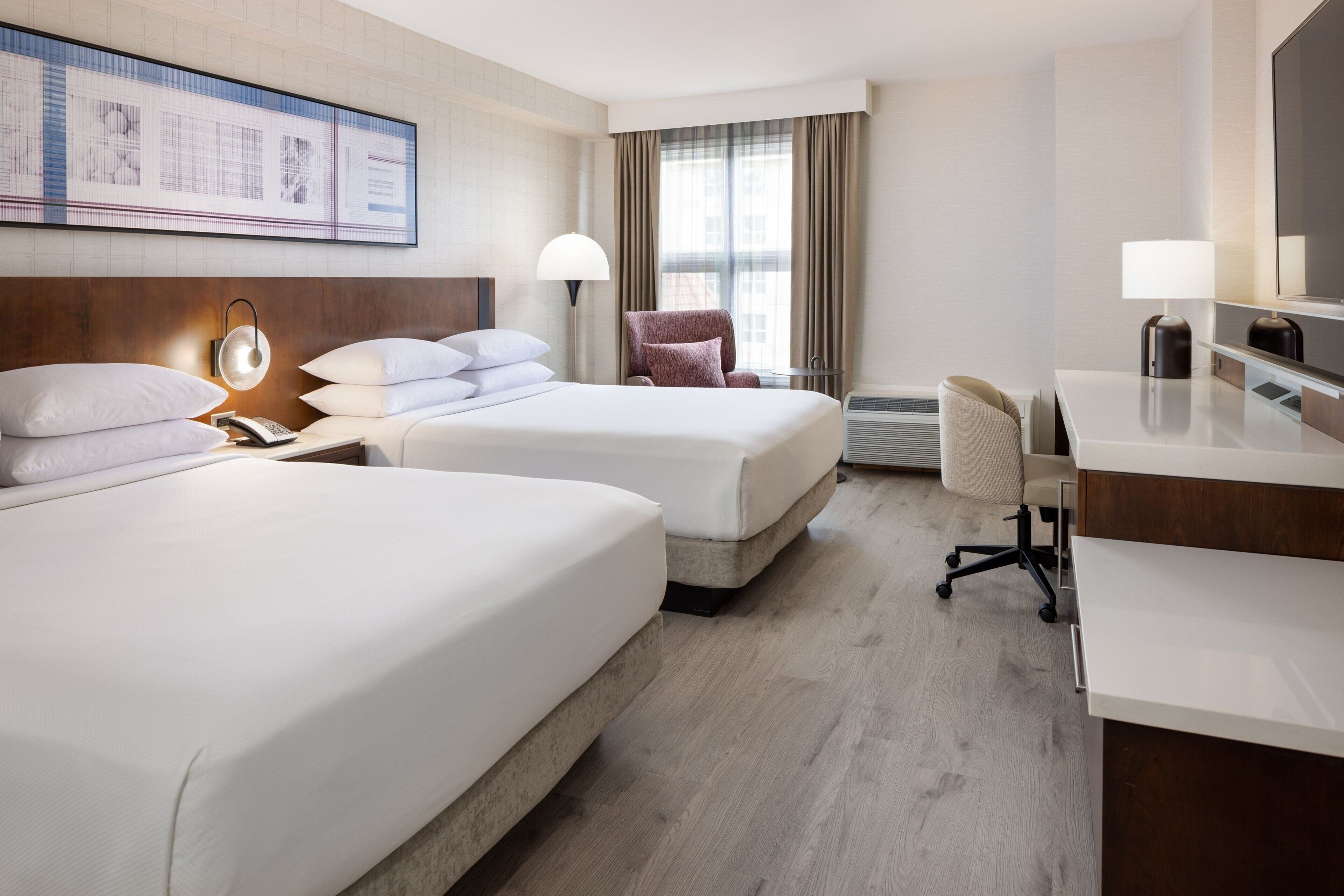 A hotel room with two beds, a desk, a chair, a lamp, and a large window with curtains.