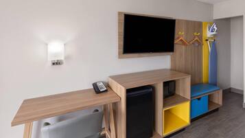 A modern hotel room with a wooden desk, a flat-screen TV, a mini-fridge, and a colorful storage unit.