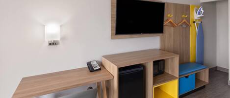 A modern hotel room with a wooden desk, a flat-screen TV, a mini-fridge, and a colorful storage unit.