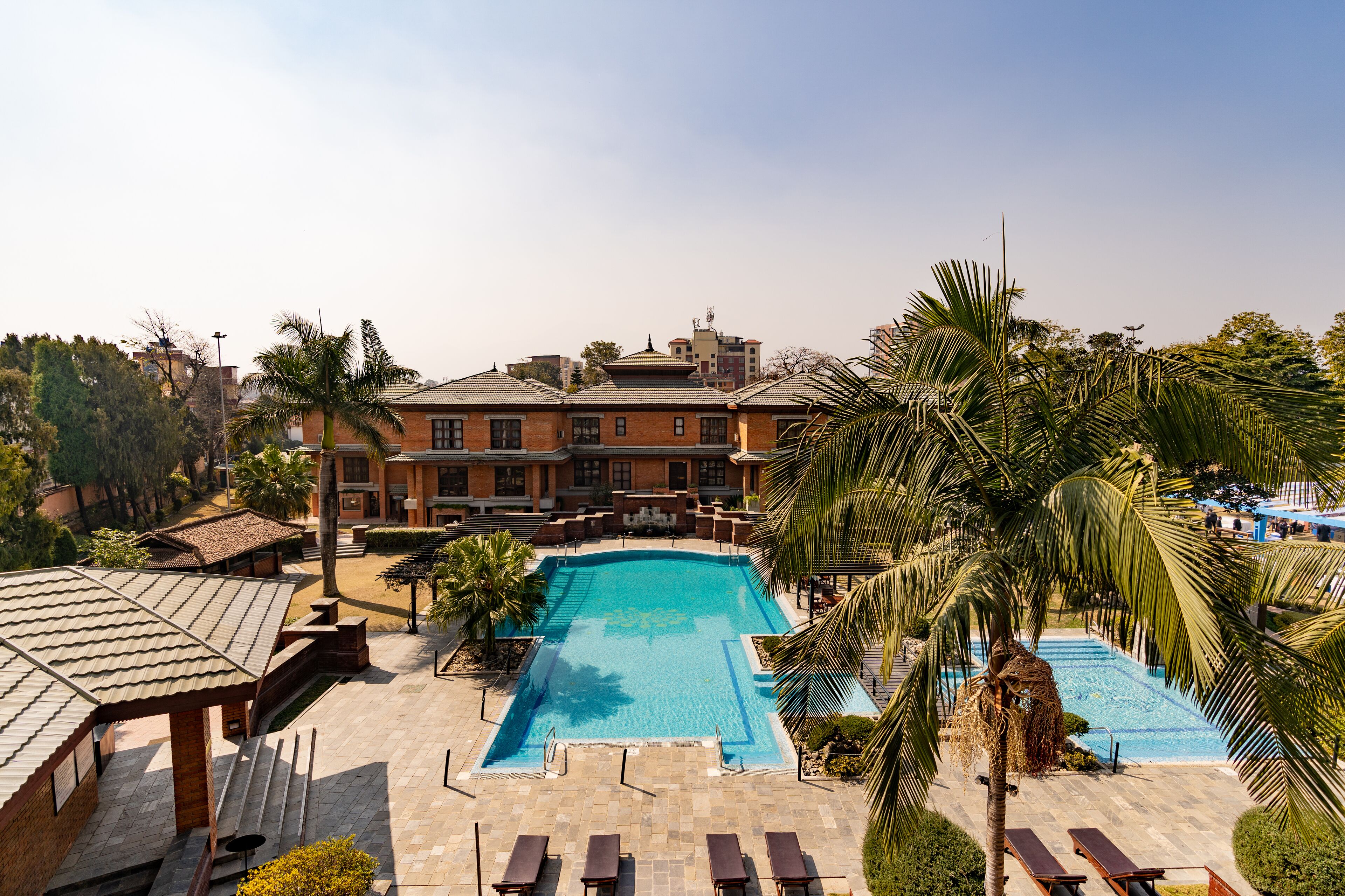 Una piscina amplia con zona de deck adoquinada, rodeada de palmeras y sillones de descanso.