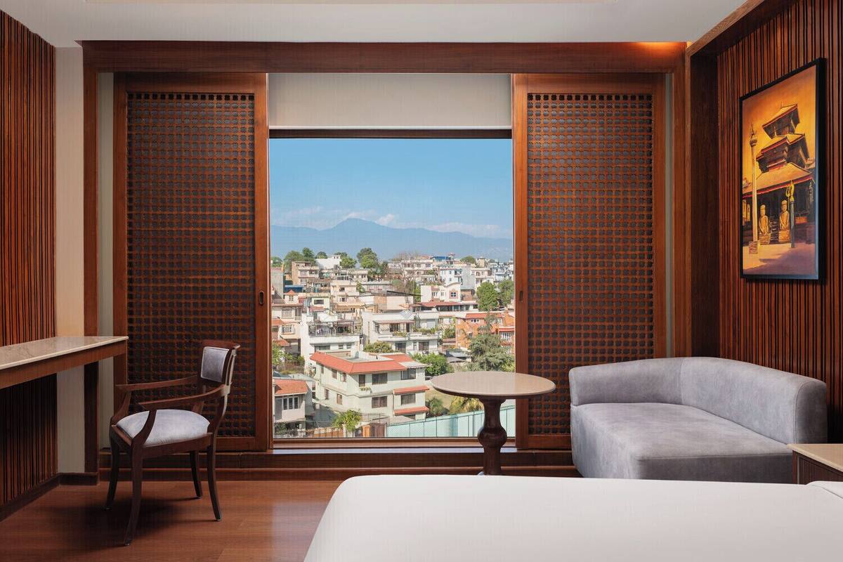 A hotel room with a bed, a chair, a small table, and a view of a cityscape through a large window.