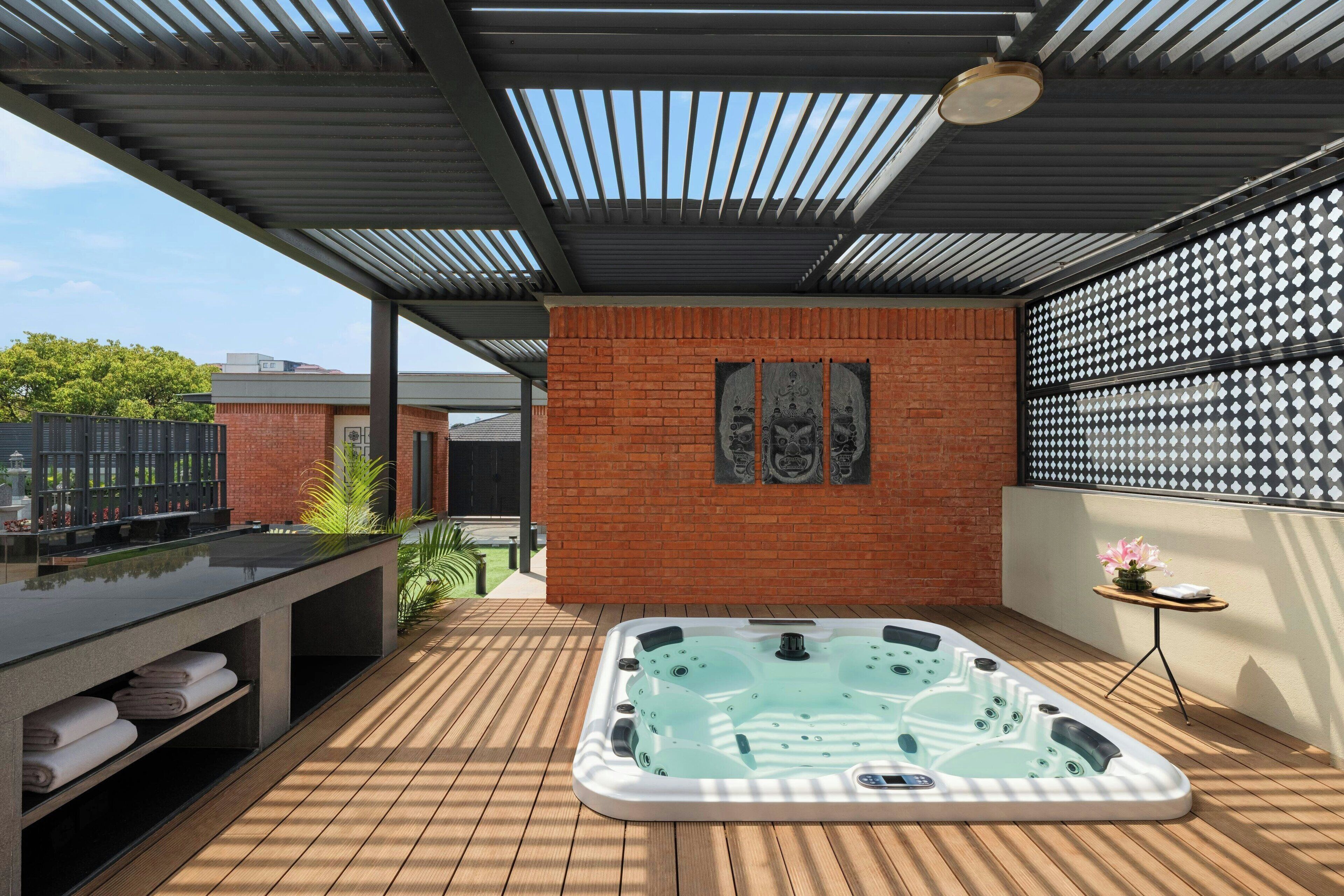 A modern outdoor hot tub area with a wooden deck, a brick wall, and a pergola overhead.
