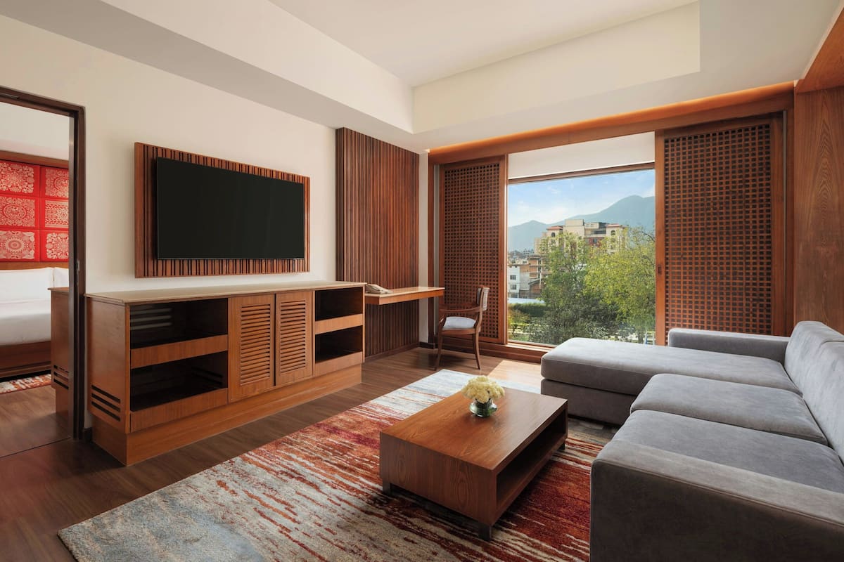 A modern living room with a large flat-screen TV, wooden furniture, a grey sofa, and a view of the city through large windows.
