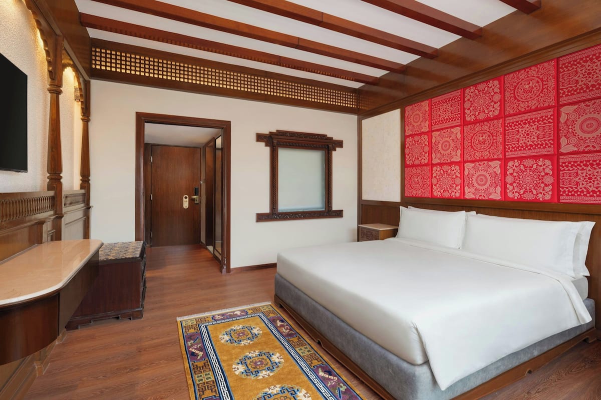 A hotel room with a wooden ceiling, a large bed with white bedding, a wooden desk, and a red patterned wall.