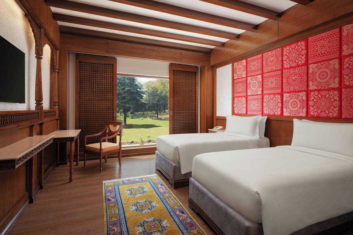 A hotel room with two beds, a desk, a chair, and a view of a garden through a sliding door.