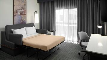 A hotel room with a bed, a desk, a chair, a lamp, and a large window with curtains.