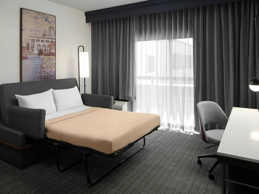 A hotel room with a bed, a desk, a chair, a lamp, and a large window with curtains.