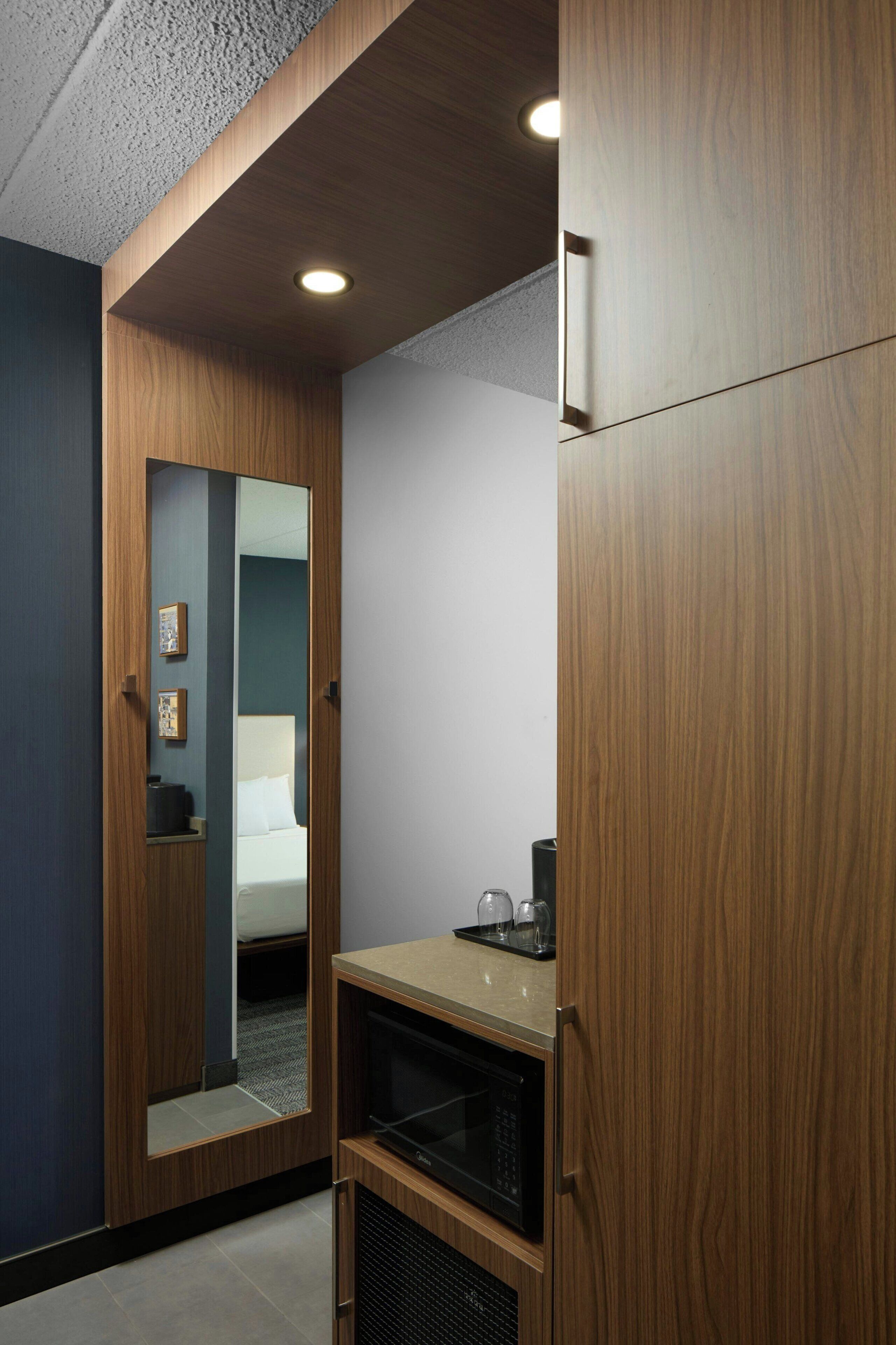 Ein kleines Hotelzimmer mit einem Holzschrank, einer Kochnische und einem Spiegel, der das Bett reflektiert.